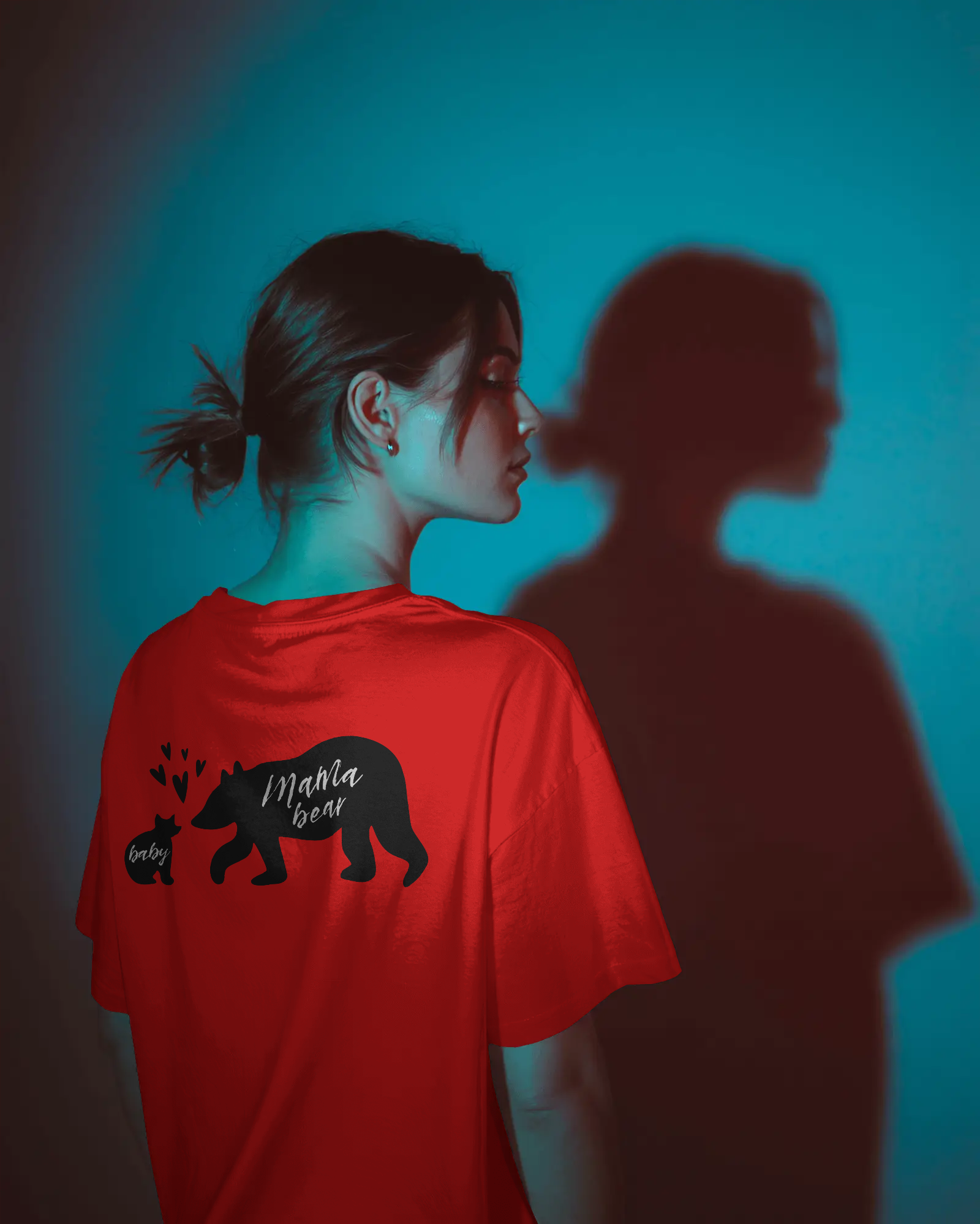 A woman wearing a red oversized T-shirt with a black graphic print of a mama bear and baby bear with hearts, standing in a creative lighting setup with a dramatic shadow on the wall.
