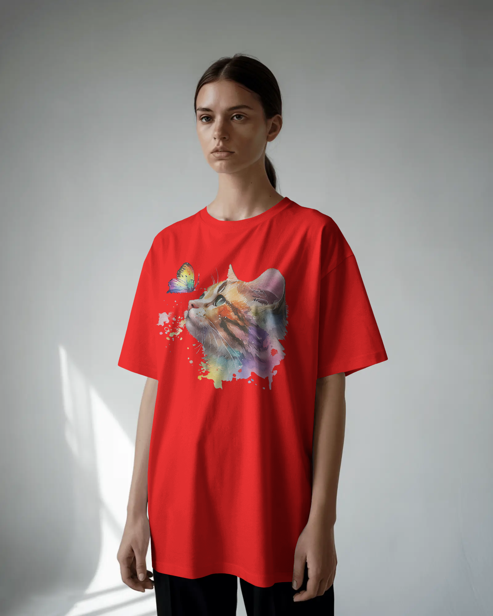 A person wearing a red oversized T-shirt featuring a colorful watercolor cat looking at a butterfly, standing in a studio with blue-green lighting.