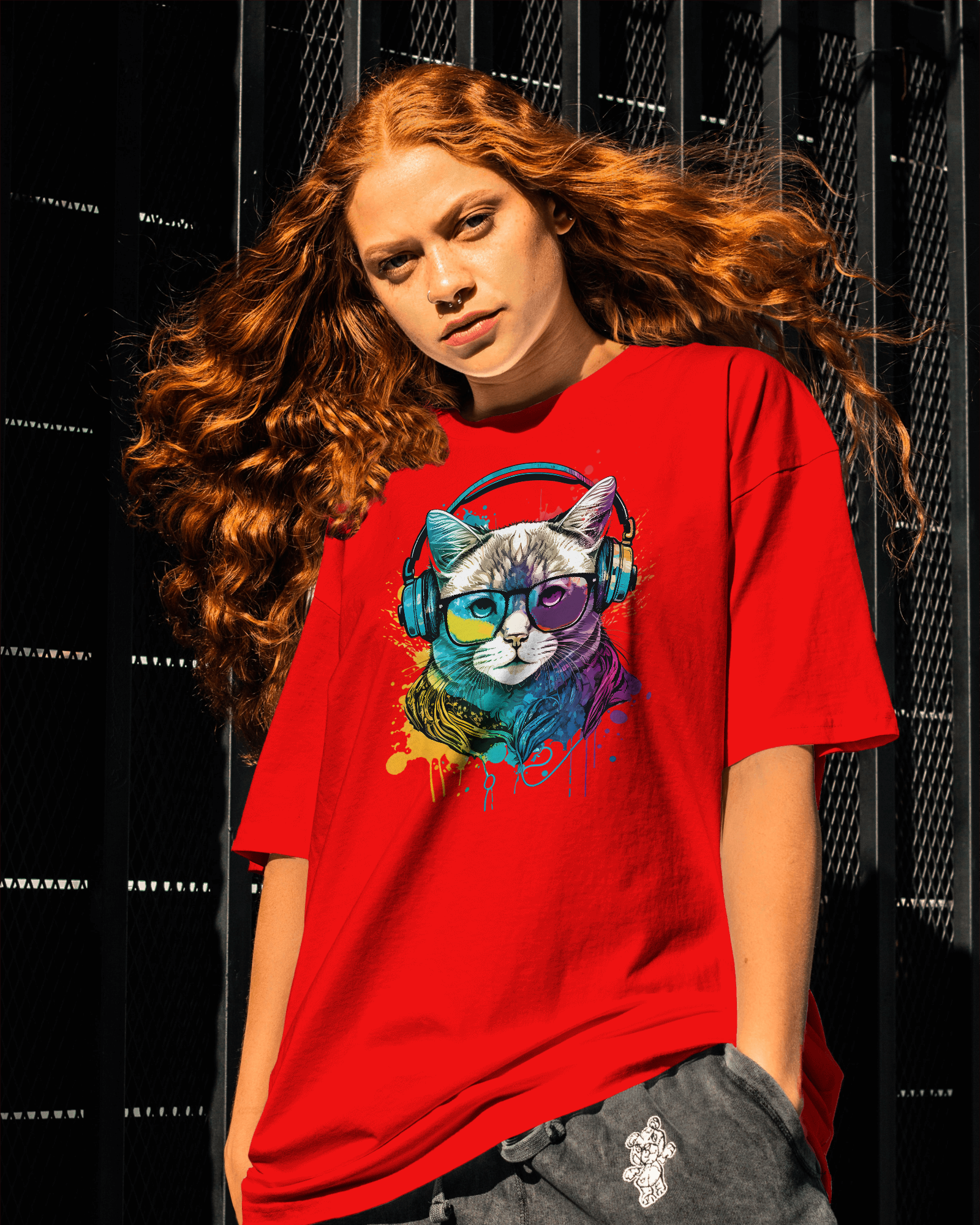 A woman wearing a red oversized T-shirt featuring a colorful neon DJ cat graphic with headphones, standing in an urban setting with metal background.