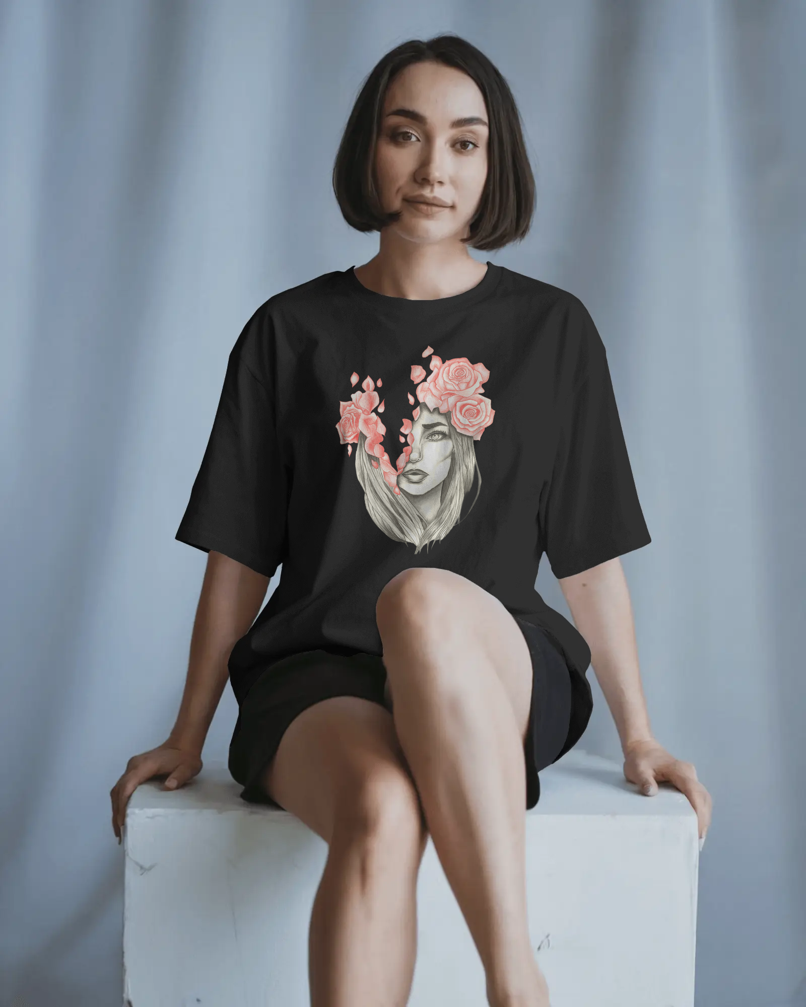 Person wearing a black t-shirt with a graphic design of a face and flowers, sitting on a white box against a gray background.