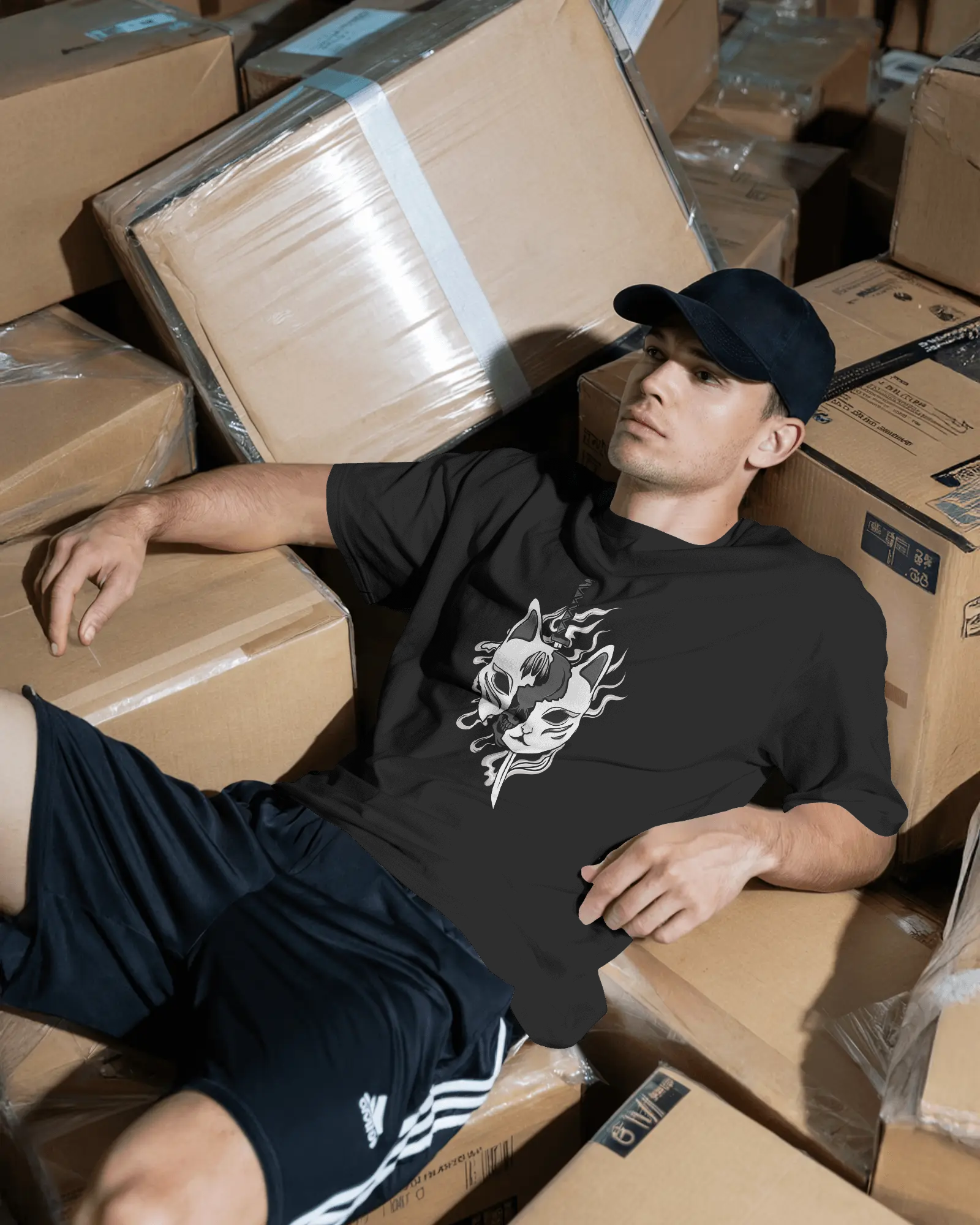 Man sitting among stacked cardboard boxes wearing a black t-shirt with a graphic design.