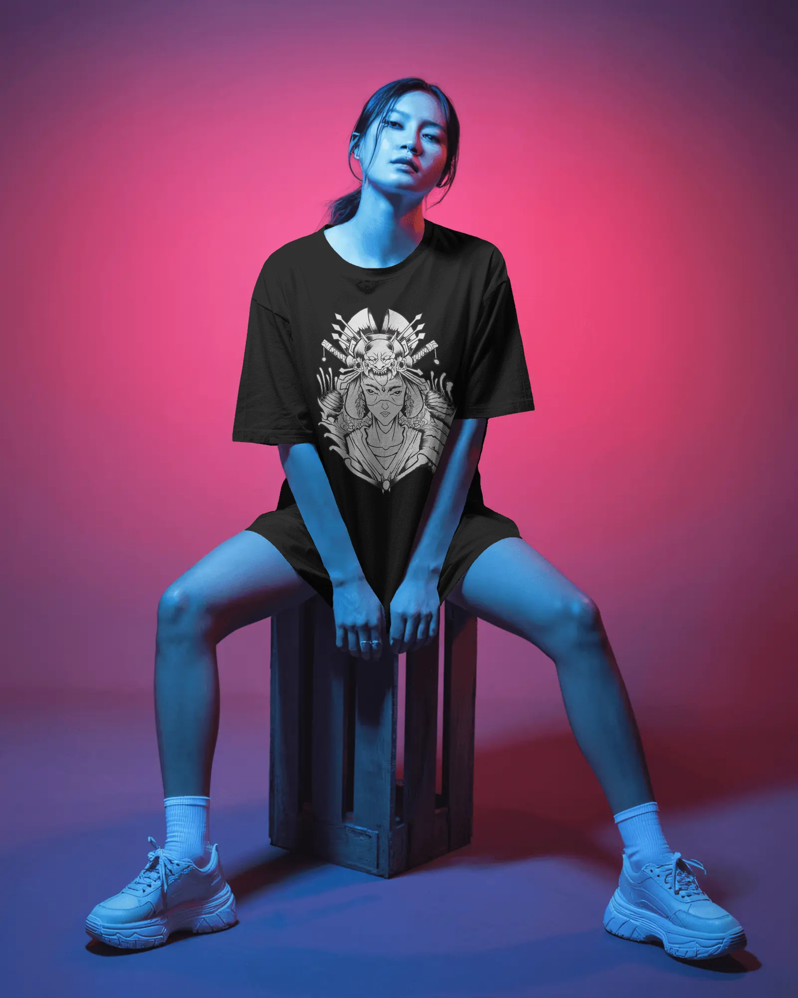 Person wearing a black t-shirt with a graphic design, sitting on a crate against a pink and purple gradient background.