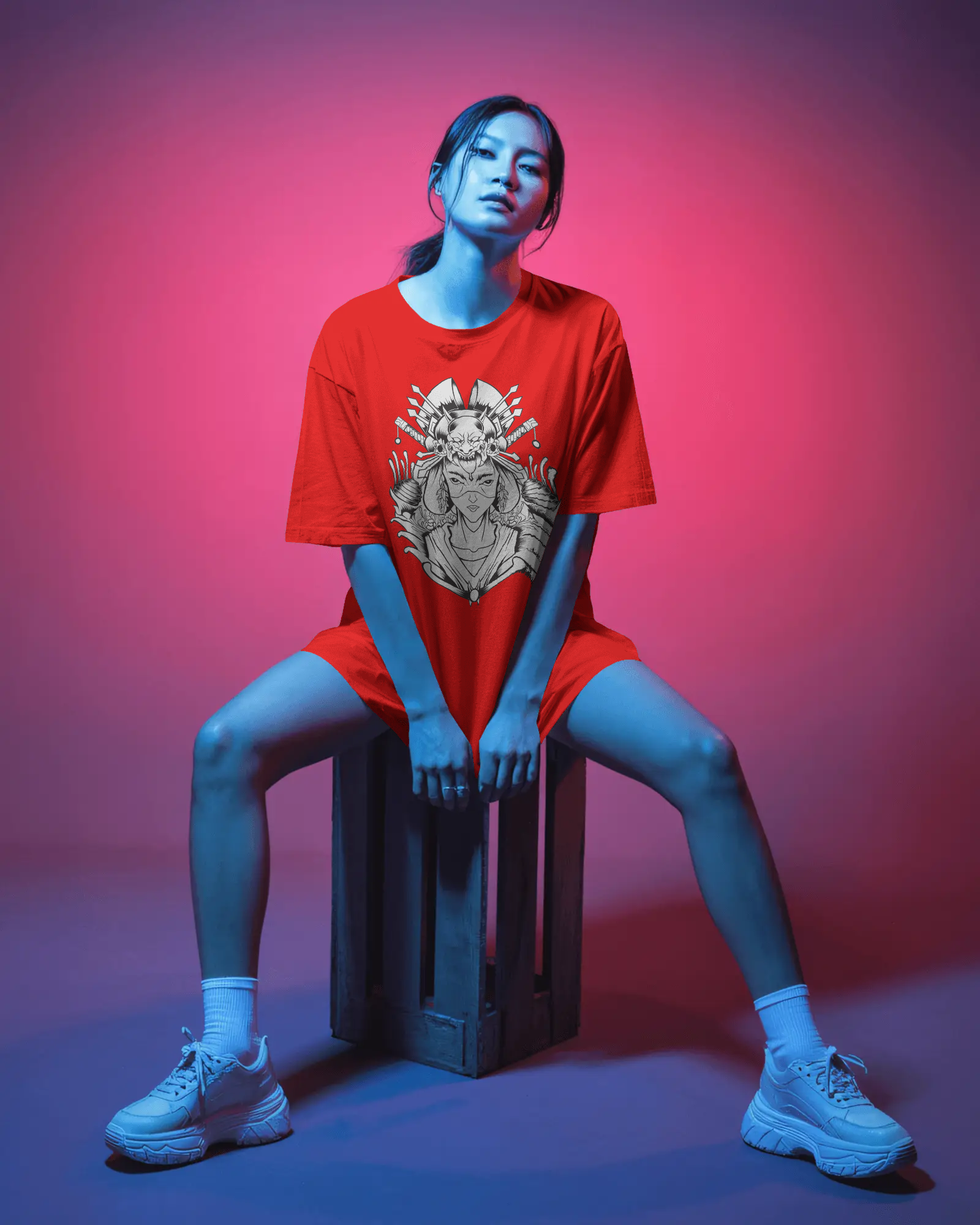 Person wearing a red t-shirt with a graphic design, sitting on a crate against a pink and purple gradient background.