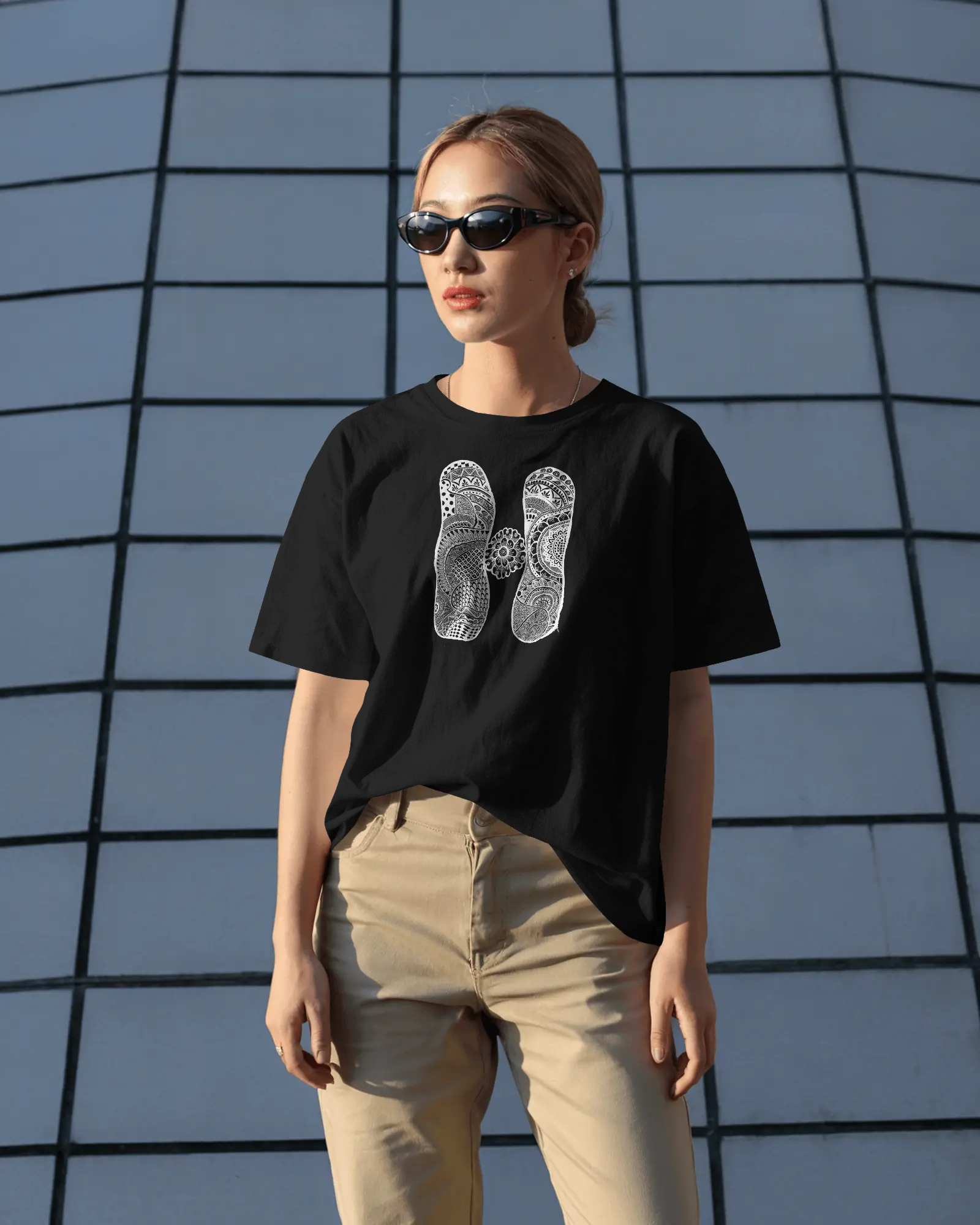 A woman wearing a black oversized T-shirt with a white artistic mandala letter H printed on the front, standing outdoors in sunlight with sunglasses.
