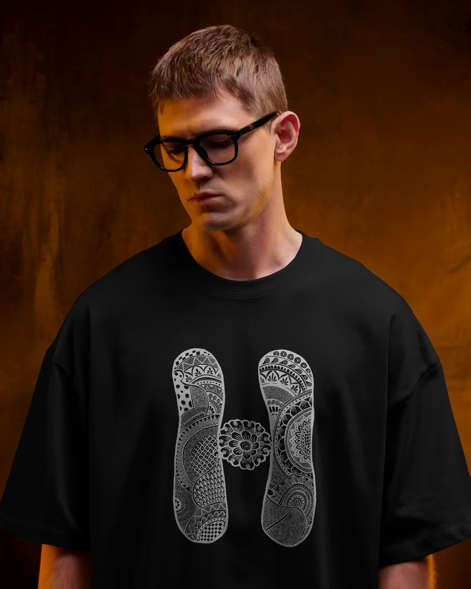 A woman wearing black oversized T-shirt with a white artistic mandala letter H printed on the front, standing outdoors in sunlight with sunglasses.