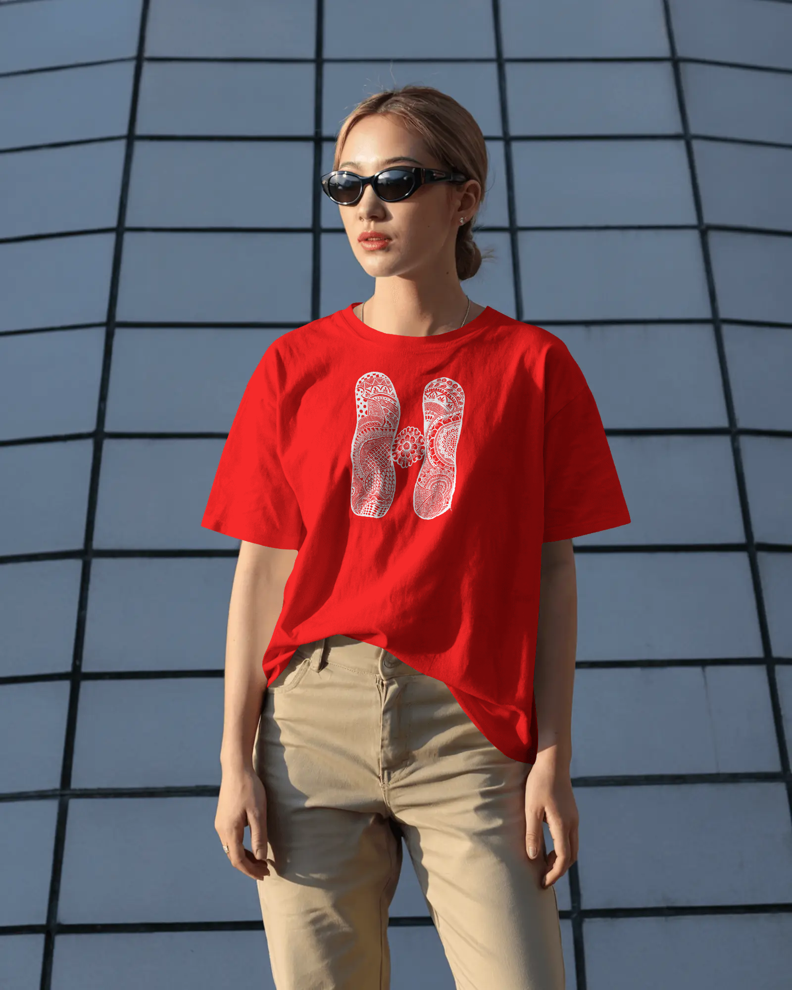 A woman wearing a red oversized T-shirt with a white artistic mandala letter H printed on the front, standing outdoors in sunlight with sunglasses.