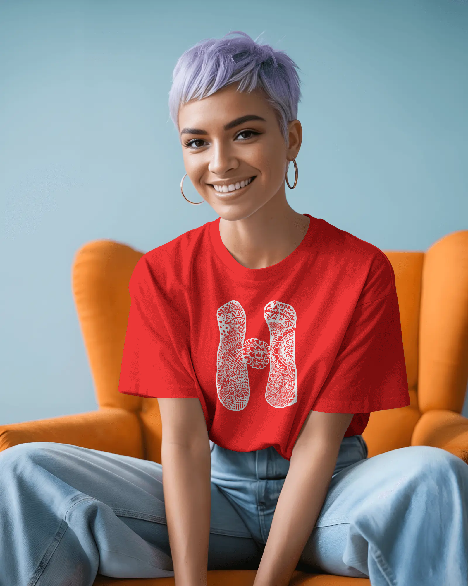 A woman wearing a red oversized T-shirt with a white artistic mandala letter H printed on the front, standing outdoors in sunlight with sunglasses.