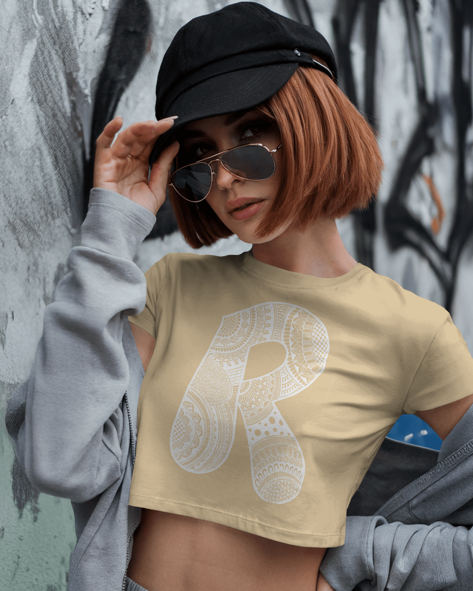 Person wearing a beige t-shirt with a large letter 'R' graphic, black cap, and sunglasses against a graffiti-covered wall.