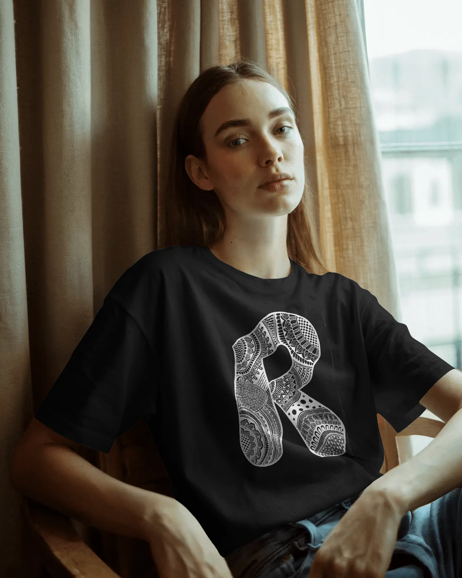 A woman wearing a black oversized T-shirt with a detailed white mandala letter R printed on the front, standing against a light neutral background.