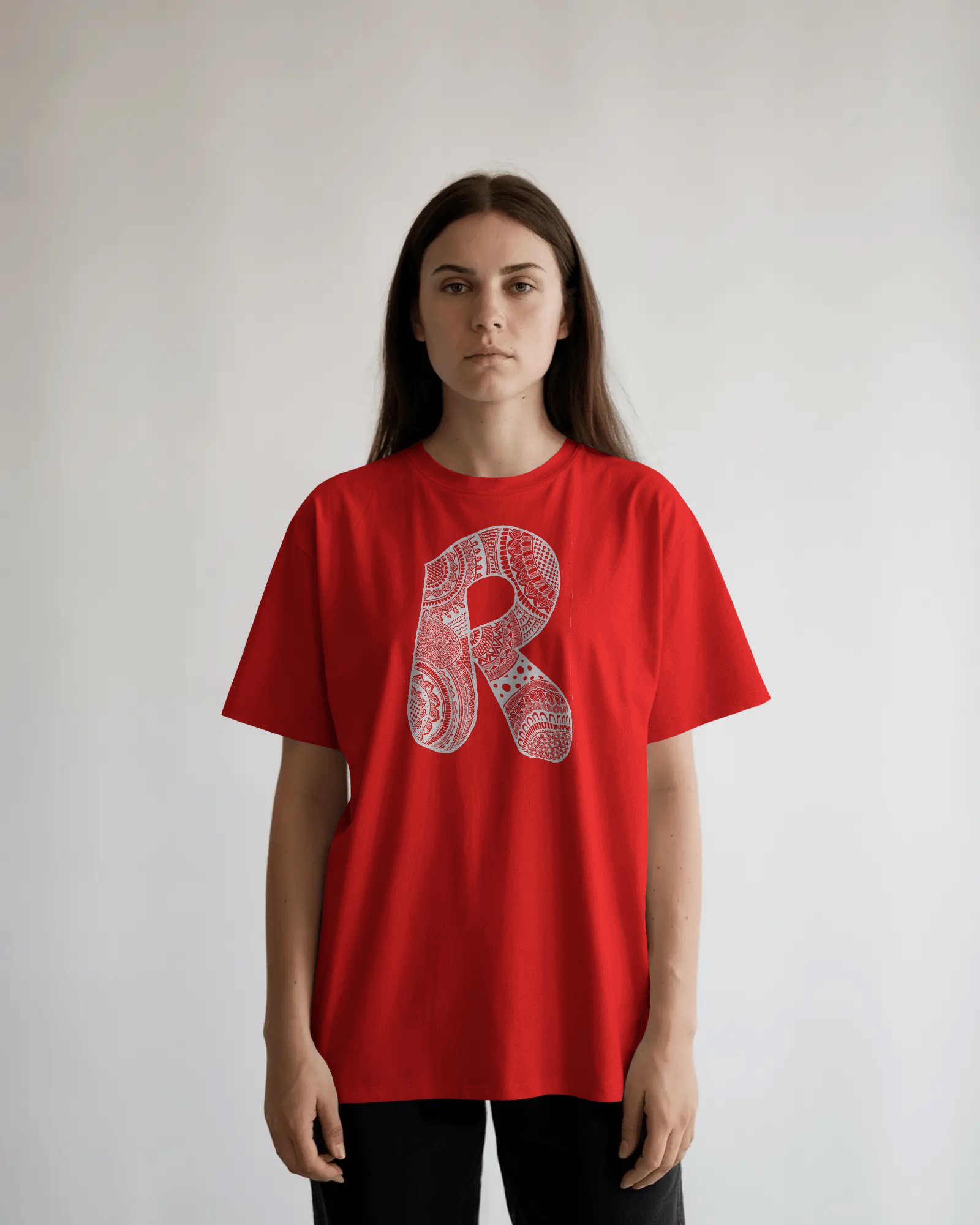 A woman wearing a black oversized T-shirt with a detailed white mandala letter R printed on the front, standing against a light neutral background.