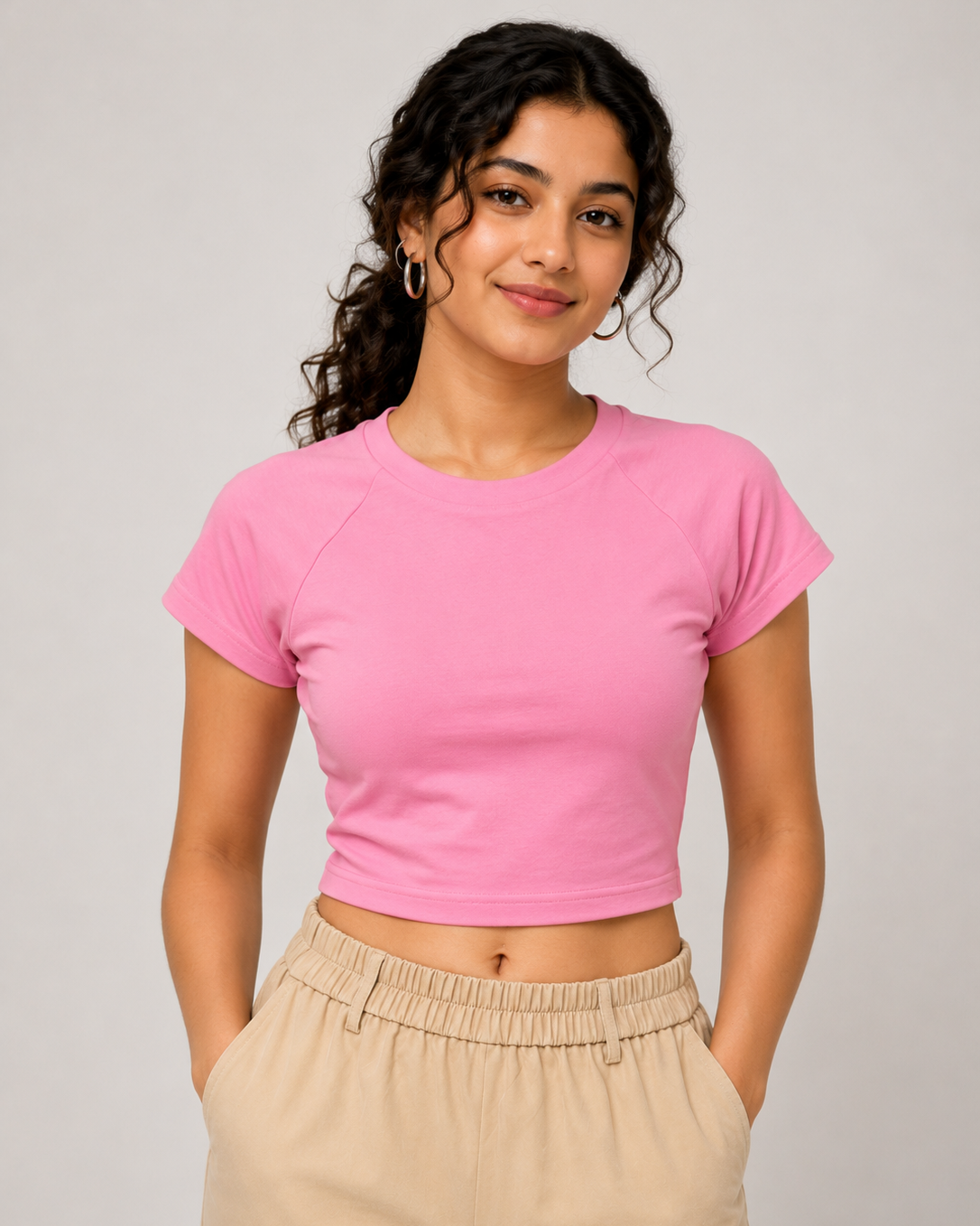 A woman modeling a bright pink short-sleeve crop top with raglan sleeves, paired with beige drawstring pants, against a solid light gray background.