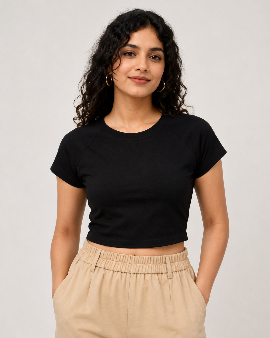 A woman with curly dark hair wearing a black short-sleeve crop top and beige high-waisted trousers, standing against a light gray studio background.