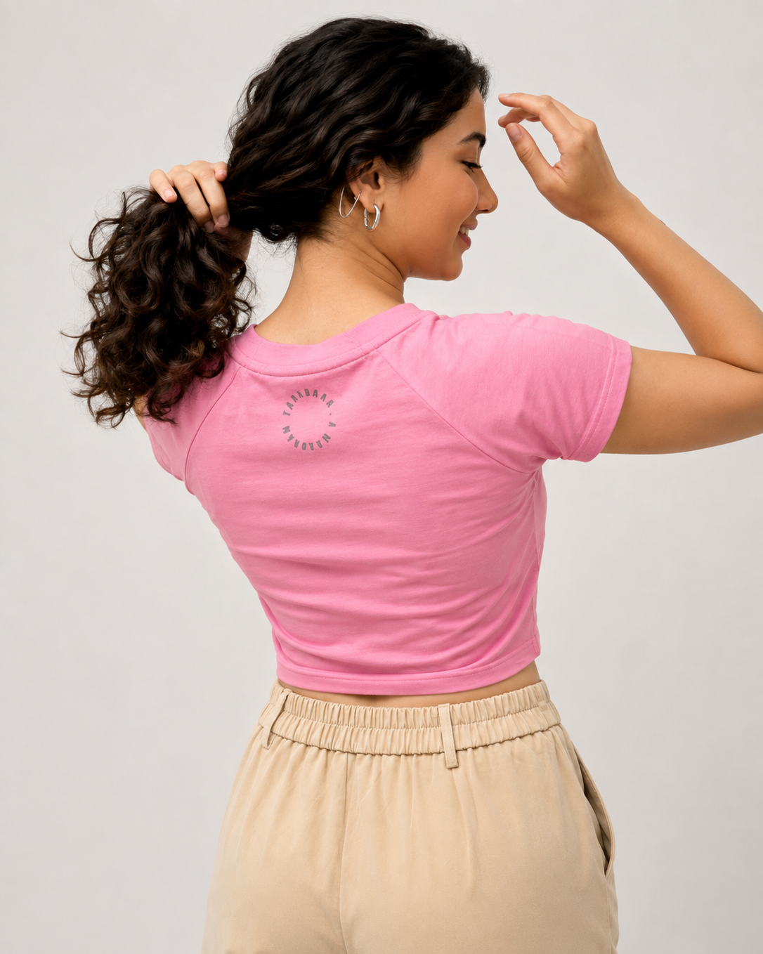 A woman shown from the back wearing a pink short-sleeve crop top and beige drawstring pants against a light gray studio background.