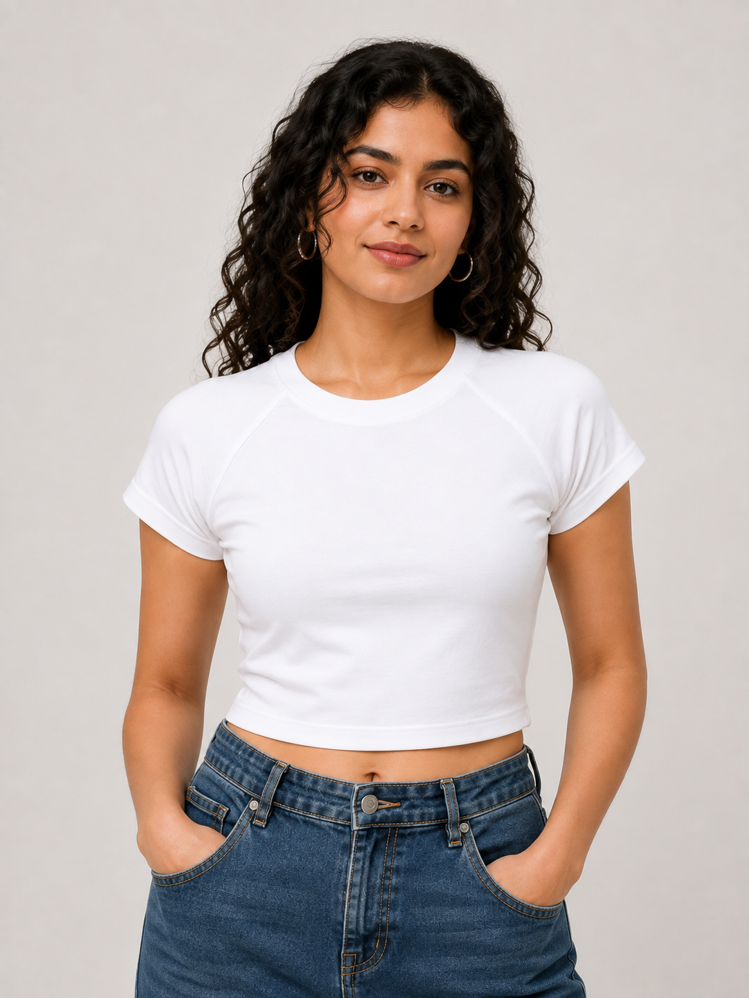 A woman with curly dark hair wearing a white short-sleeve crop top and blue jeans, standing against a light gray studio background.