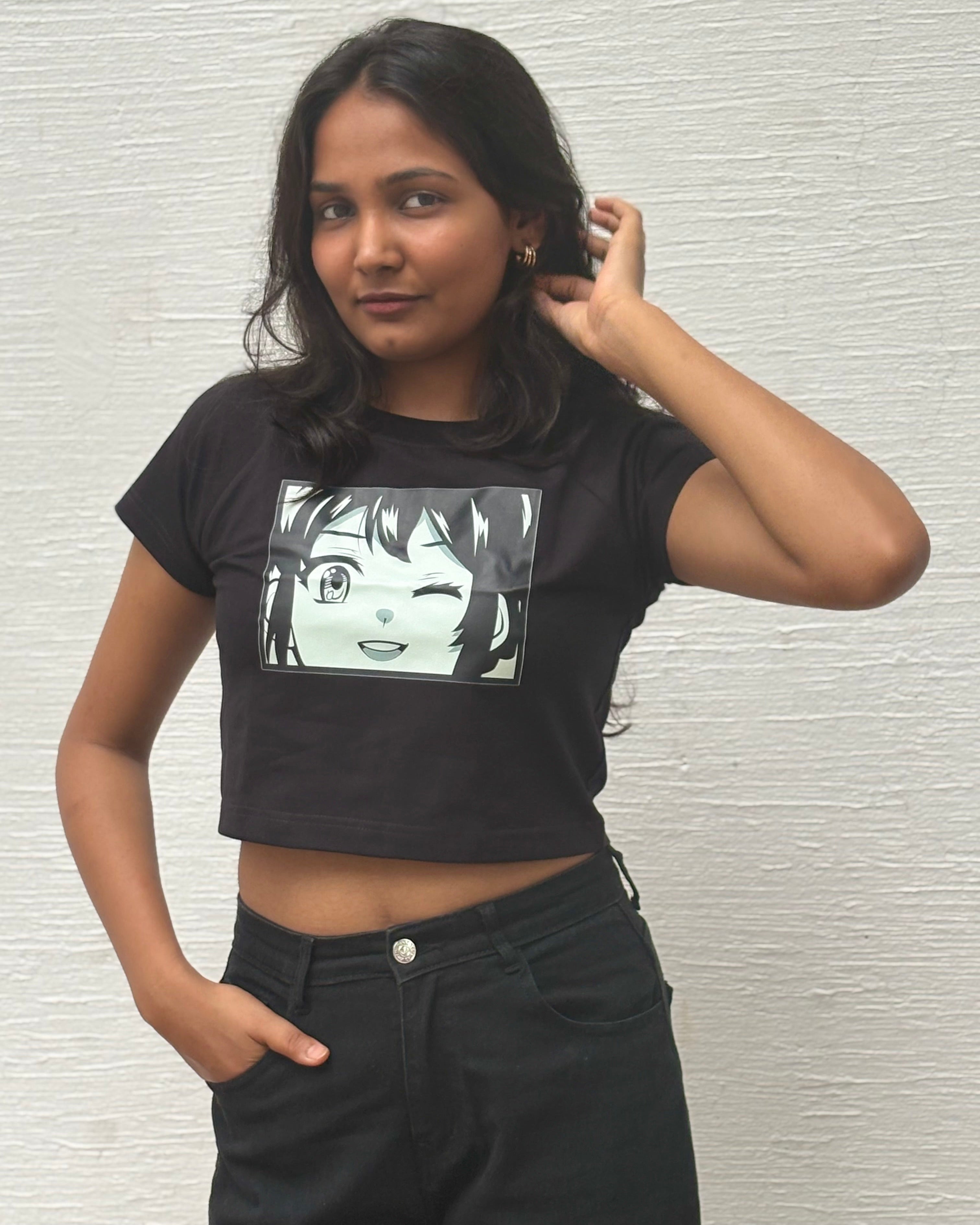 A woman wearing a black short-sleeve nazar na lage crop top t-shirt with a black and white anime girl graphic on the front, paired with black jeans, standing against a white textured wall.