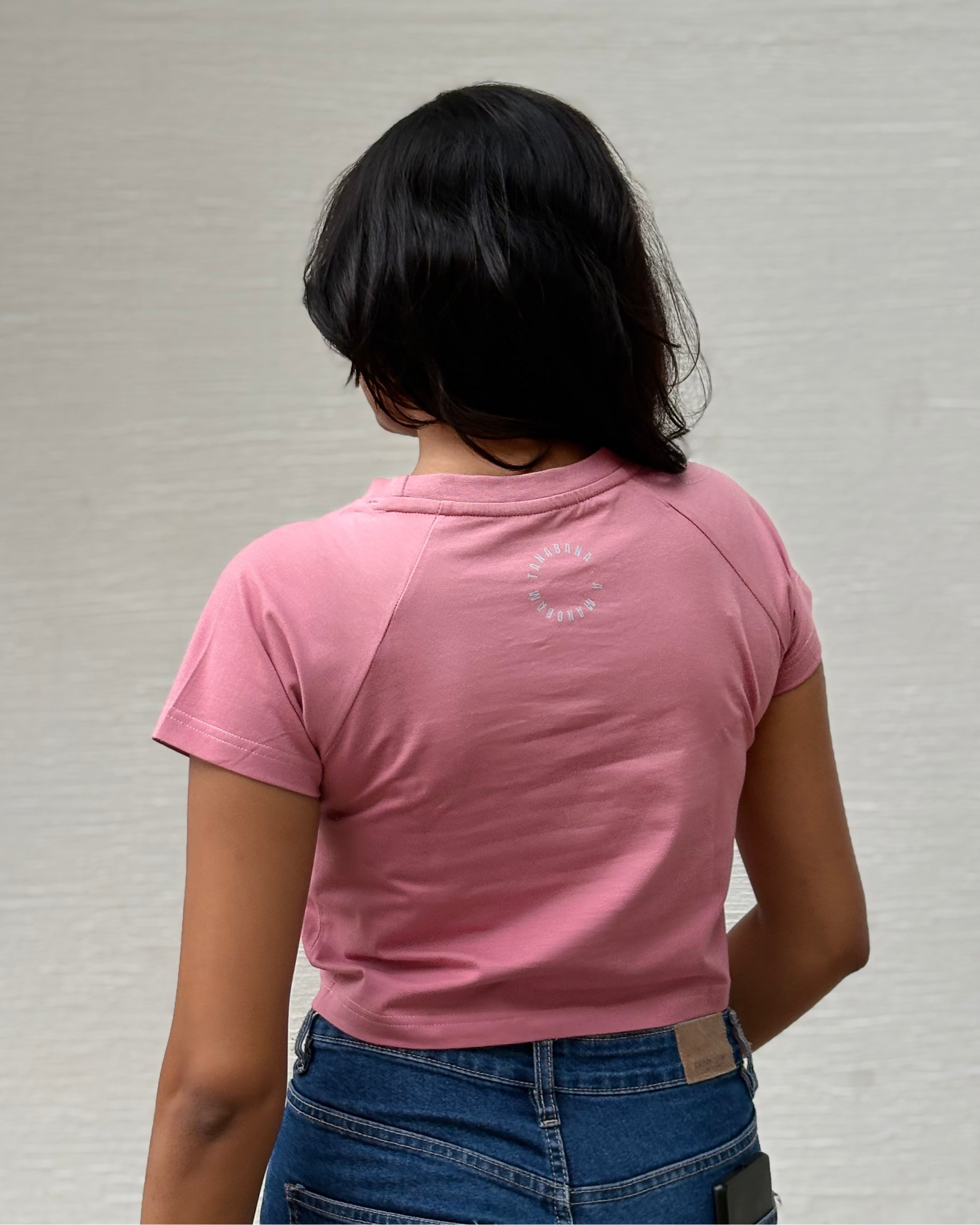 A person viewed from behind wearing a pink short-sleeve t-shirt and blue jeans, standing against a textured off-white wall.