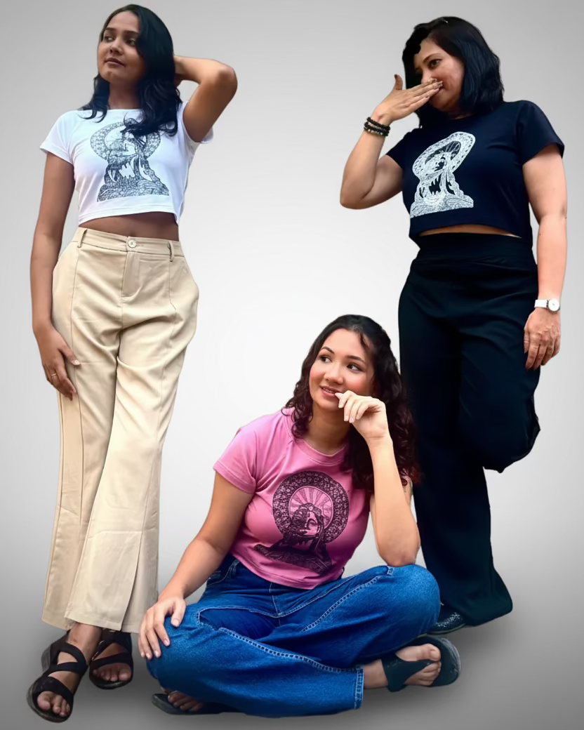 Three women posing together outdoors against a textured concrete wall with green vines. The woman on the left wears a white graphic crop top and beige wide-leg pants.