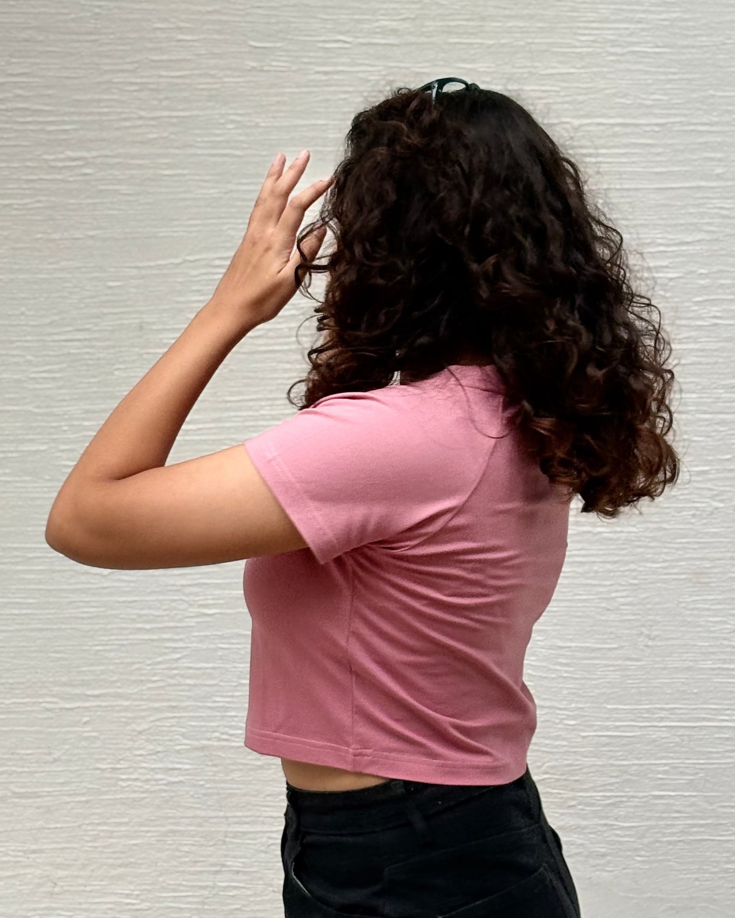 A person with long, curly dark brown hair is shown from behind, wearing a pink short-sleeve crop top and black pants, posing against a textured off-white wall.